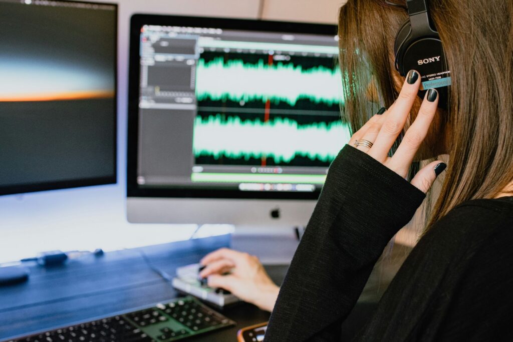 A woman wearing a black long-sleeve shirt and Sony headphones using a computer to work on audio editing software.