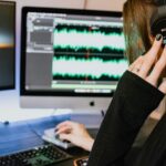 A woman wearing a black long-sleeve shirt and Sony headphones using a computer to work on audio editing software.