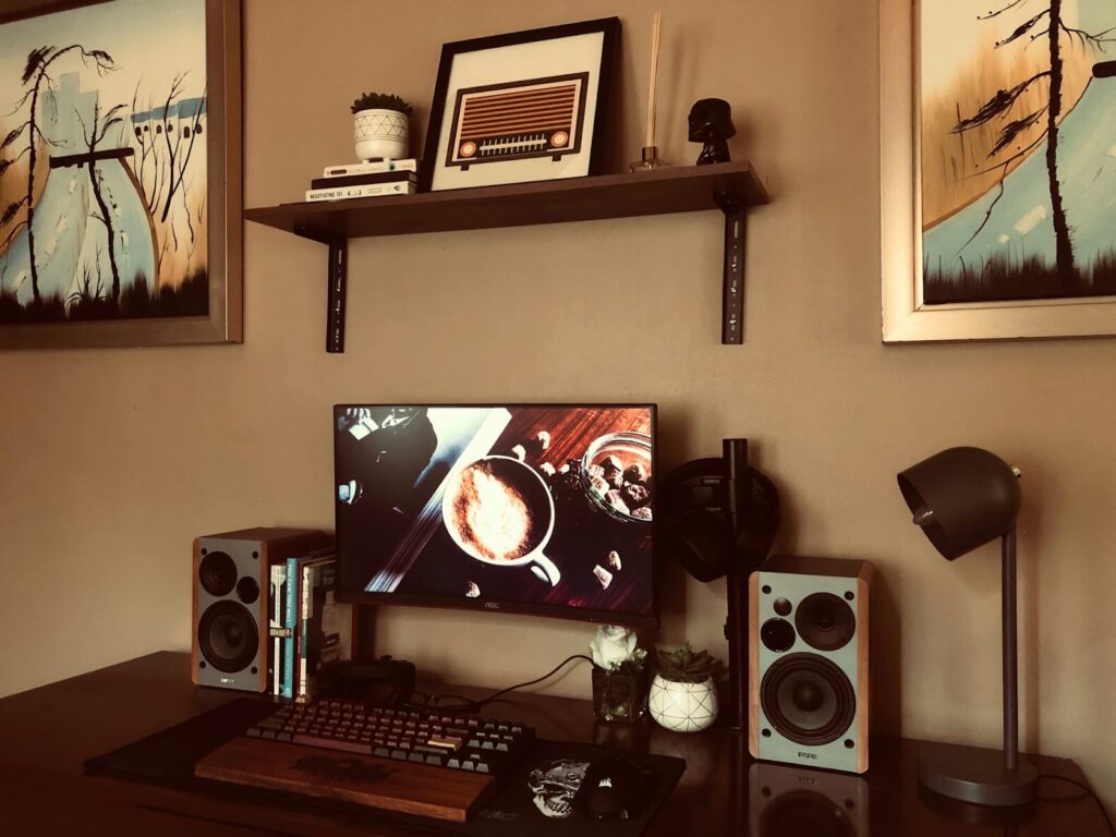 Computer desk setup with monitor, keyboard, and speakers. A decorative shelf and wall art hang overhead.